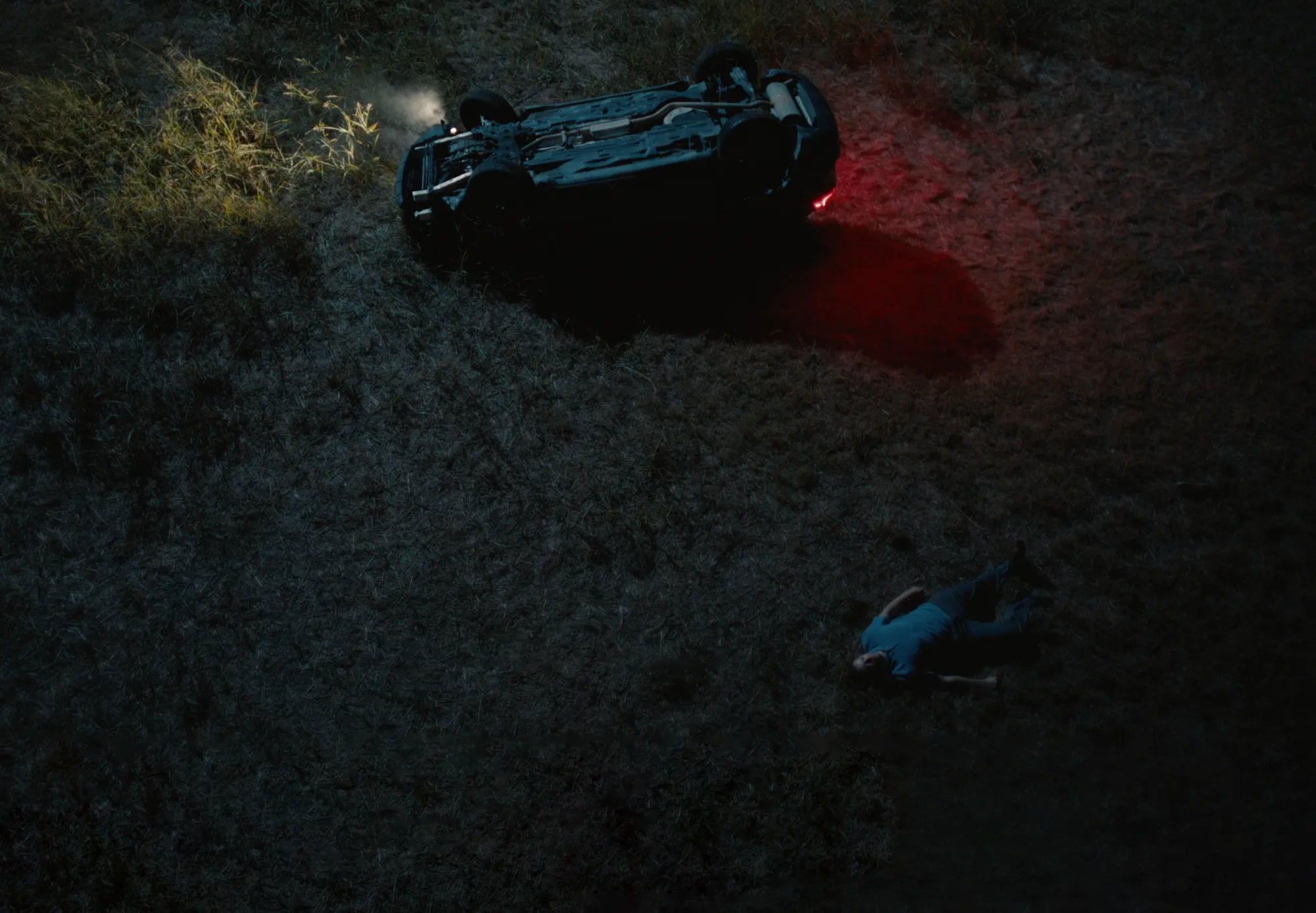 Overturned car with rear lights on and smoke coming from it, next to a person lying on the ground in grass at night.