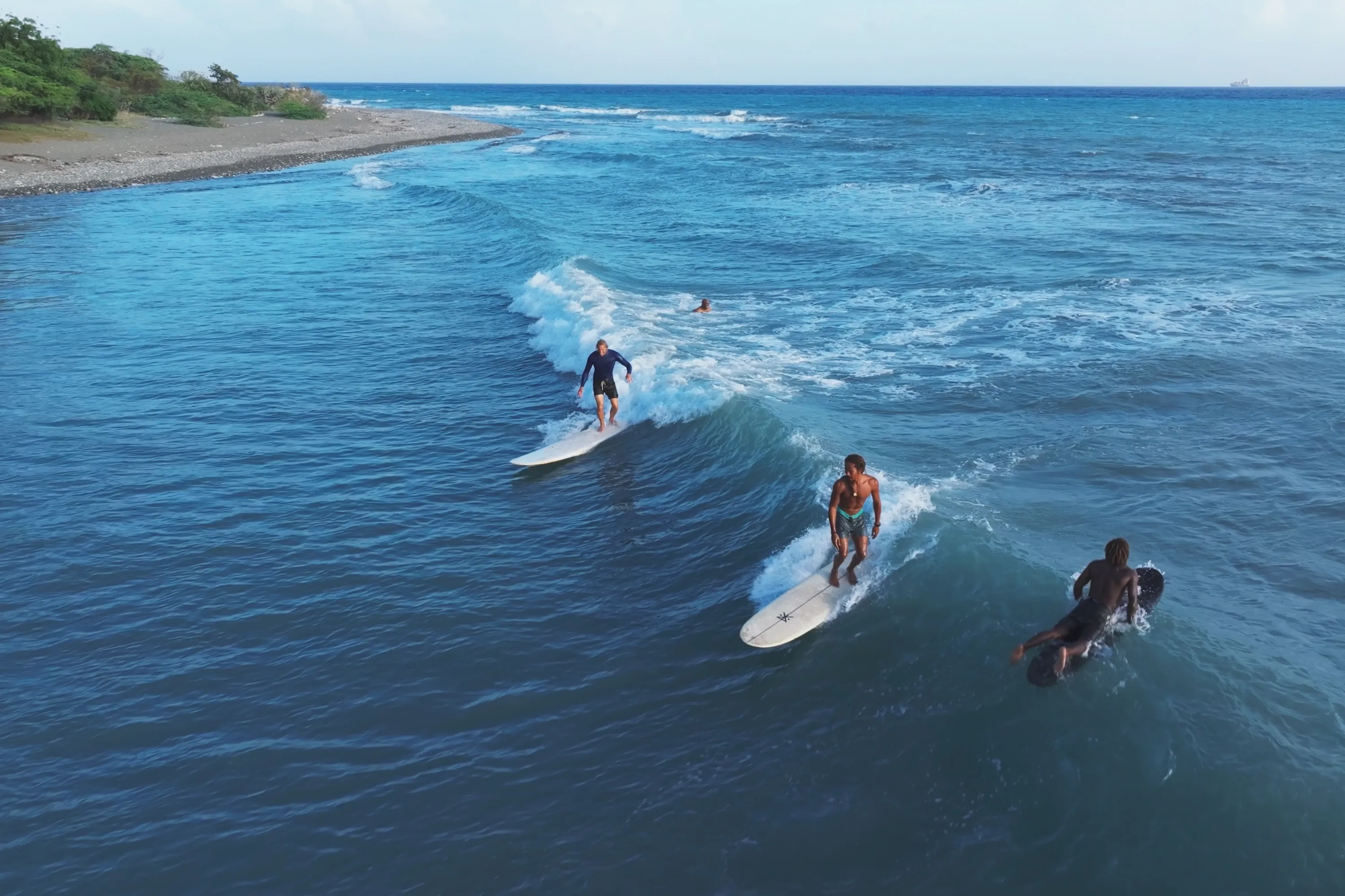 Three surfers riding ocean waves near a rocky shoreline with greenery under a clear sky.